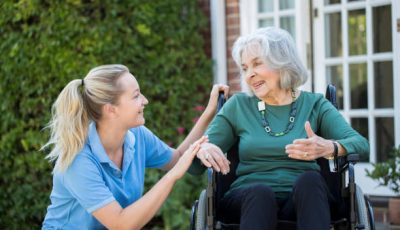 Carer Pushing Senior Woman In Wheelchair Outside Home